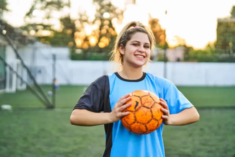 A confident, smiling teenage soccer player holds a soccer ball in her hands