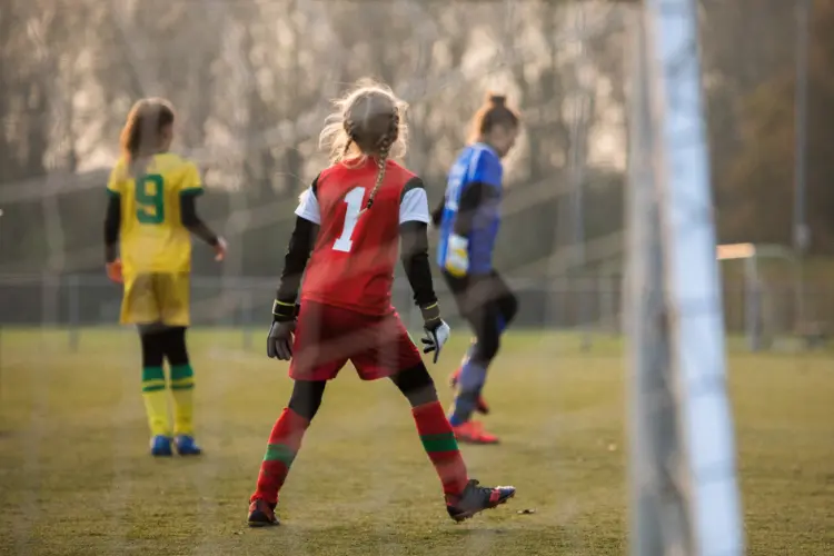 A youth goalkeeper watches the ball at the other end of the field and stays ready and on her toes