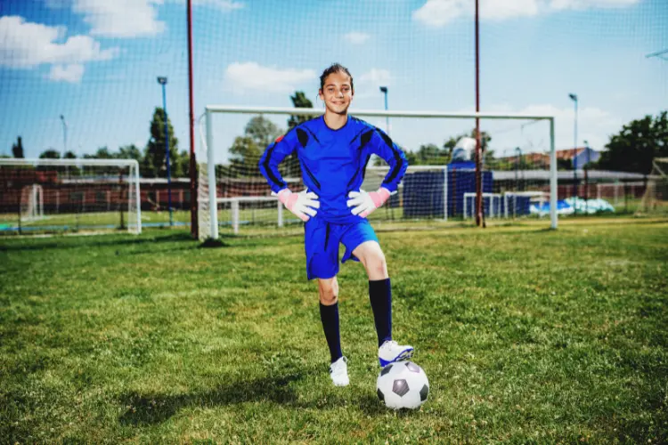 A confident goalkeeper poses for a photo with her hands on her hips and her left foot on the soccer ball