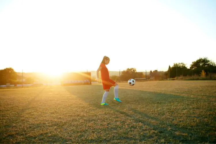 A girl practices her juggling skills on a grass field at sunset while wearing a red uniform and teal cleats