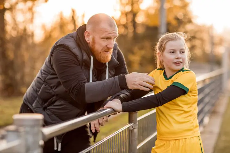 Father talks with his daughter while leaning on a railing during soccer practice