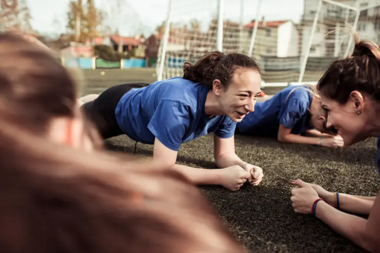 Teen girls do planks together on a soccer field while laughing and smiling