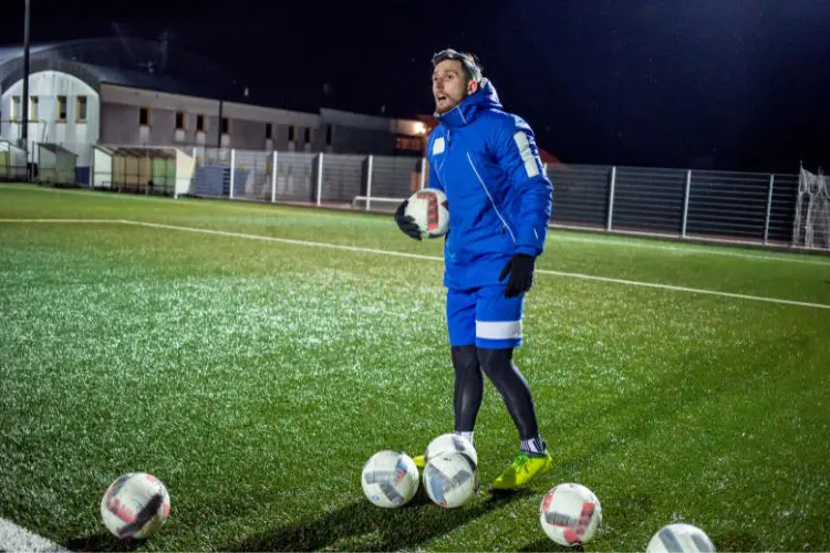 A soccer coach stands with a pile of balls and yells instructions to his team