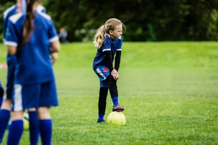 A girl leans on a soccer ball while listening to instructions from her coach