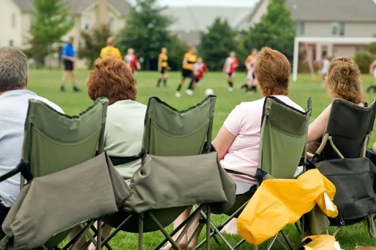 A line of soccer parents sit on the sideline at a game