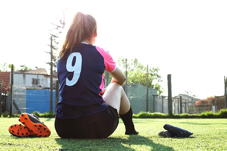 Soccer player sits looking at the field with her cleats and shinguards in a pile next to her