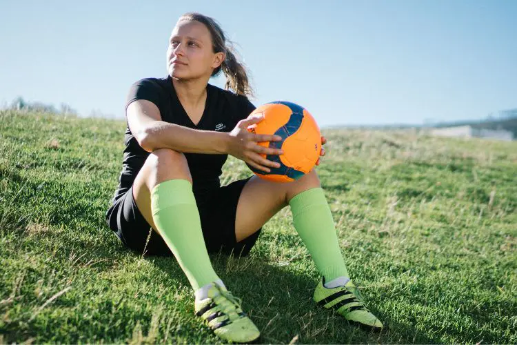 A women's soccer player holds a soccer ball in her hands while sitting on a hill