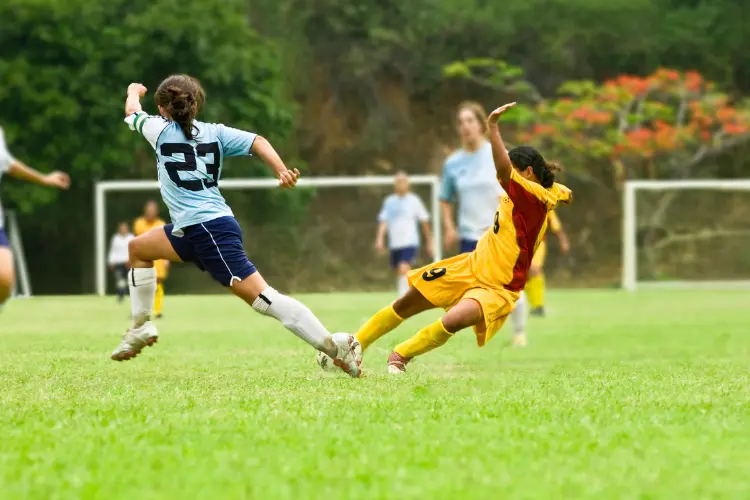 Soccer player striking the ball as she falls to the ground while another player runs in front to block the ball