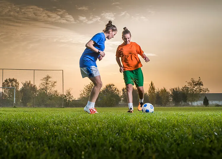 Two girls playing pickup soccer 1v1 on a grass field at sunset