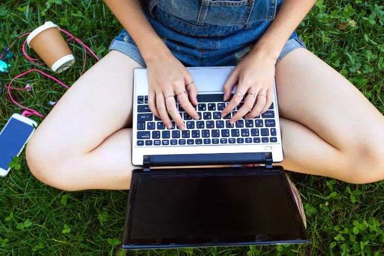 A girl in overalls sits in the grass using a laptop with coffee and headphones